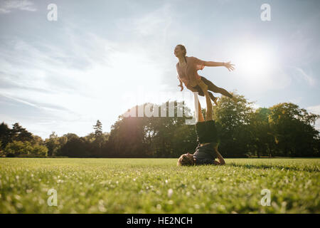 Fit young couple doing acrobatic yoga in park. Man lying on grass and balancing woman in his feet. Stock Photo