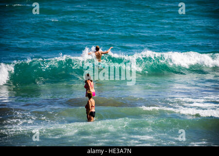 Two middle aged women paddle in the sea while watching a young man jump through the surf Stock Photo