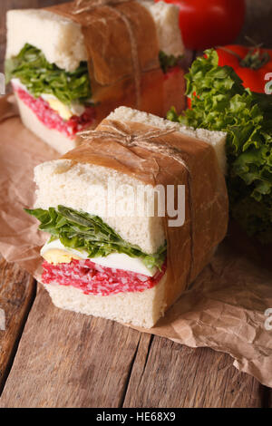 rustic sandwiches with salami, lettuce and egg wrapped in paper close-up on the table. vertical Stock Photo