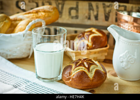 Fresh lye rolls in a wicker basket, a glass of milk, white pitcher on a wood table in a rustic style kitchen interior Stock Photo