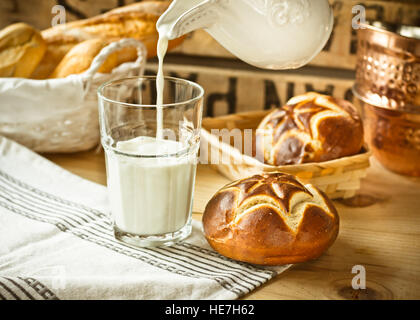 Fresh lye rolls in a wicker basket, process of pouring milk into a glass from pitcher, wood table, rustic style kitchen interior Stock Photo