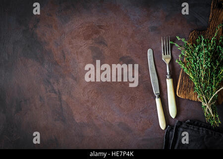 Vintage silverware with ivory handle, dark textile, wooden cutting board and bunch of fresh thyme on dark rusty background. Top Stock Photo