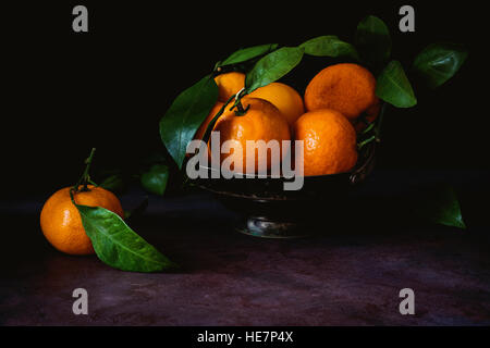 Still life of fresh tangerines with green leaves in vintage metal bowl. Toned image, low key technique (chiaroscuro). Close up Stock Photo