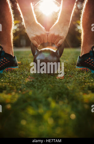 Cropped shot of young man doing kettlebell workout, focus on hands holding kettle bell on grass. Stock Photo