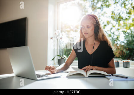 Young female student studying at home. Young woman sitting at table with book using laptop. Stock Photo