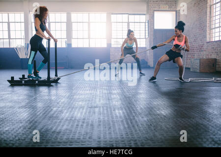 Shot of woman standing on sled with other pulling in gym. Three young females doing physical training at healthclub. Stock Photo