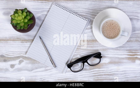 Notebook and pen with reading glasses, plant and creamy coffee on rustic white table Stock Photo