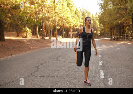 Cute young brunette jogger stretching in a forest on a tarred road cutting through it with bright sunshine from behind her Stock Photo
