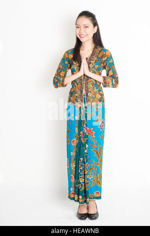 Portrait of young southeast Asian woman in traditional Malay batik kebaya dress greeting, full length standing on plain background. Stock Photo