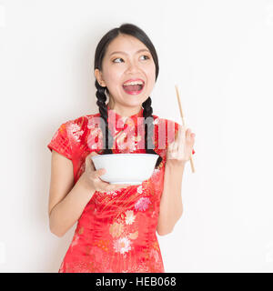 Portrait of young Asian woman in traditional cheongsam dress eating, hand holding bowl and chopsticks, celebrating Chinese Lunar New Year or spring fe Stock Photo
