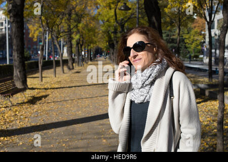 happy woman with grey cardigan and scarf, black sunglasses, calling mobile phone in urban street in Madrid city with trees in autumn Stock Photo