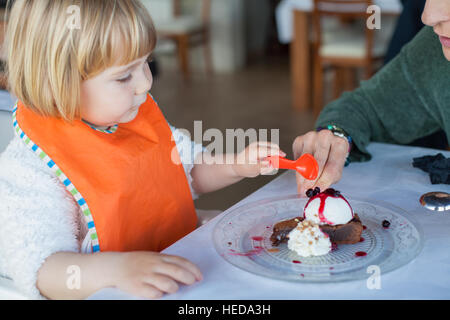 two years old child with orange plastic spoon sharing with woman a piece of chocolate cake with vanilla ice cream on crystal dish on white tablecloth Stock Photo