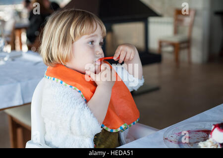 two years old child eating with orange plastic spoon and bib a piece of chocolate cake with vanilla ice cream at restaurant Stock Photo