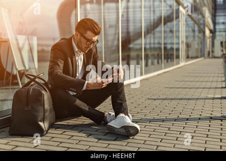 Handsome bearded businessman in the airport station typing text message on his mobile phone and sitting on the ground next to entrance Stock Photo