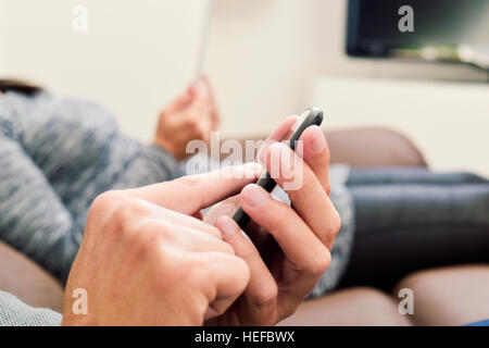 closeup of a young caucasian man sitting on a couch using his smartphone and a young caucasian woman next to him using her tablet Stock Photo