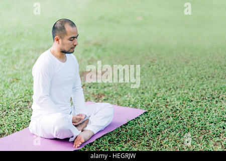 Handsome Asian man meditating on green grass in the park, calm and focused, health and yoga meditation concept, with copy space Stock Photo