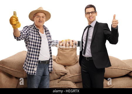 Elderly farmer and a businessman giving thumbs up in front of a pile of burlap sacks filled with potatoes isolated on white background Stock Photo