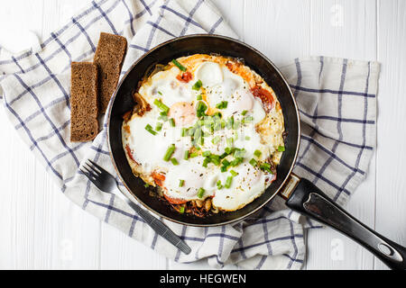 scrambled eggs with tomatoes and basilic in black frying pan on white wood background Stock Photo