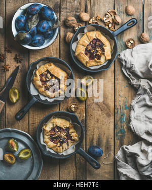 Plum and walnut crostata pie with ice-cream scoops in individual cast iron pans over rustic wooden table, top view, copy space, vertical composition. Stock Photo