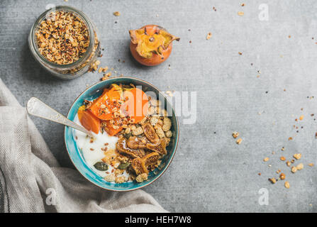 Healthy breakfast concept. Oatmeal, quinoa granola with yogurt, dried fruit, seeds, honey, persimmon in bowl over grey concrete background, top view, Stock Photo