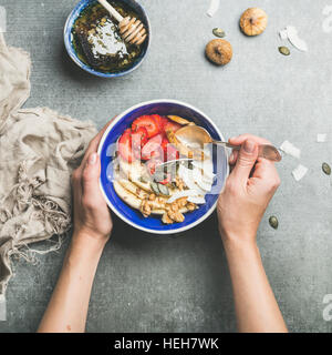 Yogurt, granola, seeds, fresh and dry fruit and honey in blue ceramic bowl in woman' s hands over grey concrete background, top view, square crop. Cle Stock Photo