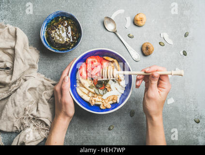 Healthy breakfast bowl. Yogurt, granola, seeds, fresh and dry fruits and honey in blue ceramic bowl in woman' s hands over grey background, top view. Stock Photo