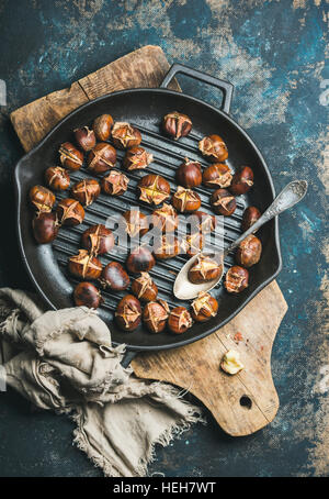 Roasted chestnuts in cast iron grilling pan over rustic wooden board and dark blue shabby plywood background, top view, vertical composition Stock Photo