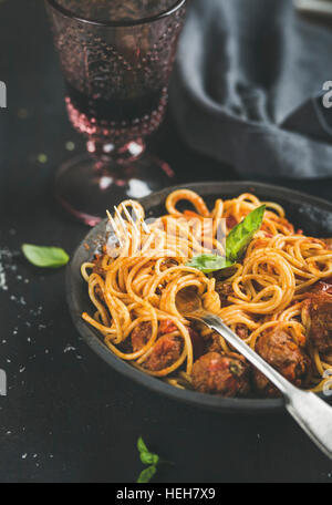 Italian pasta dinner. Spaghetti with meatballas, fresh basil leaves in dark plate and red wine in vintage glass over black background, selective focus Stock Photo