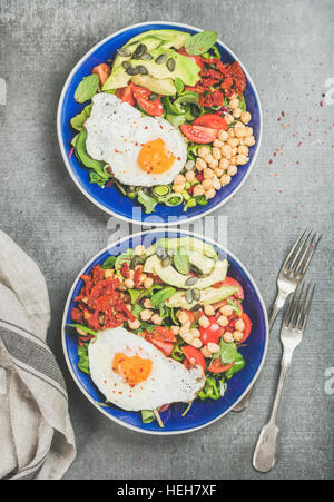 Healthy breakfast with fried egg, chickpea sprouts, seeds, fresh vegetables and greens in blue bowls over grey concrete background, top view. Clean ea