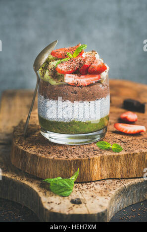 Healthy vegan breakfast concept. Matcha, almond milk, cocoa chia seed pudding with fresh fruit, berries, mint, chocolate in glass over wooden board, g Stock Photo