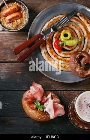 Big spiral fried sausage, meat snacks wienerwurst, ham, marinated chili peppers served in salted pretzels and plate with glass of lager beer and musta Stock Photo