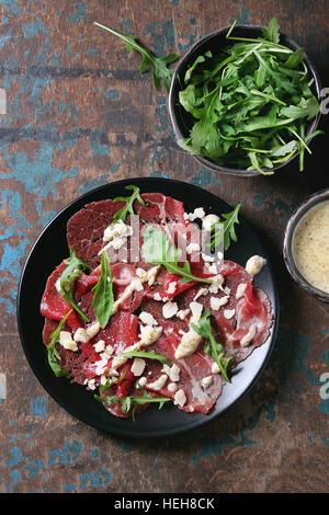 Beef carpaccio on black plate with mustard and parmesan sauce, cheese and arugula over old dark texture background. Top view with space for text Stock Photo