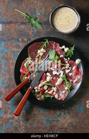 Beef carpaccio on black plate with mustard and parmesan sauce, cheese and arugula, served with fork, knife and ingredients above over old dark texture Stock Photo