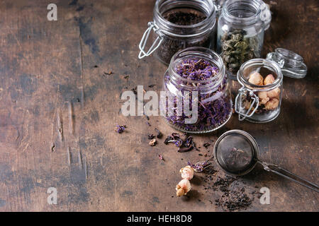Variety of black, green, rooibos, herbal dry tea leaves and rose buds in glass jars with vintage strainer over old dark wooden background. Close up wi Stock Photo
