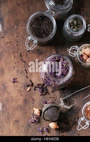 Variety of black, green, rooibos, herbal dry tea leaves and rose buds in glass jars with vintage strainer over old dark wooden background. Top view wi Stock Photo