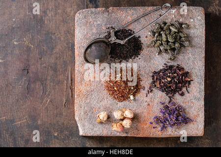 Variety of black, green, rooibos, herbal dry tea leaves and rose buds with vintage strainer on terracotta board over old dark wooden background. Top v Stock Photo