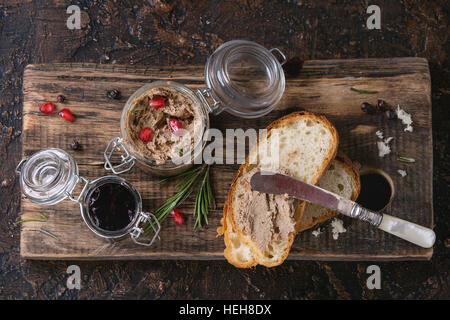 Glass jars of chicken liver pate with blackcurrant jam, pomegranate grain and sliced bread, served with vintage knife on wooden chopping board over da Stock Photo