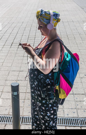 Female, high-tech tourist with a camera, backpack, headphones, and smartphone, reads as she stands on a sidewalk in Barcelona, Stock Photo