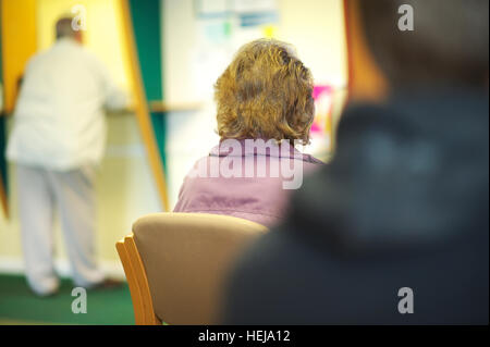 Patients sit in NHS GP waiting room while other patients stand at reception awaiting the doctor Stock Photo