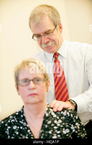 White male GP rests hand on patients back, during a back examination in his NHS clinic Stock Photo