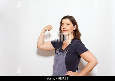 Middle aged woman in dungarees with hand on her hip and flexing the biceps muscles of the other arm, white background,copy space Stock Photo