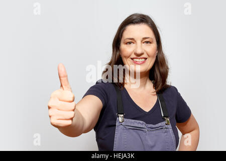 Smiling adult woman wearing dungarees showing the thumbs up sign, light grey or gray background, copy or text space Stock Photo