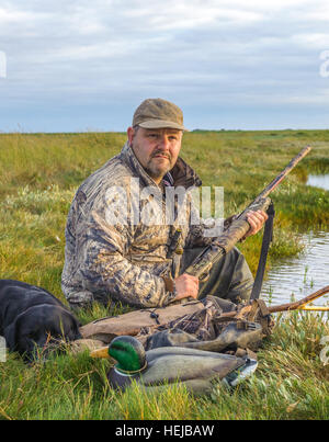 A UK wildfowler, or duck hunter, sat on the foreshore or marsh with his dog in the fading evening light Stock Photo