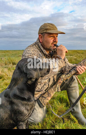 A UK wildfowler, or duck hunter, sat on the foreshore or marsh with his dog blowing a duck call in the fading evening light Stock Photo