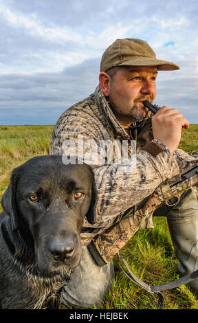 A UK wildfowler, or duck hunter, sat on the foreshore or marsh with his dog blowing a duck call in the fading evening light Stock Photo