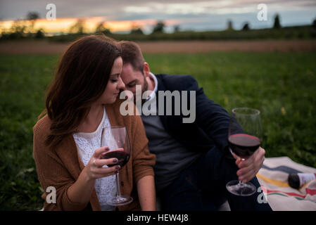 Romantic couple with red wine relaxing on picnic blanket in field at sunset Stock Photo