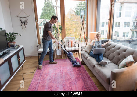 Mid adult man vacuuming with girlfriend reclining on sofa using laptop Stock Photo