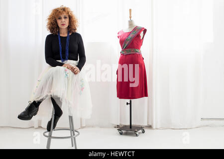 Woman sitting on stool by tailors dummy looking at camera Stock Photo