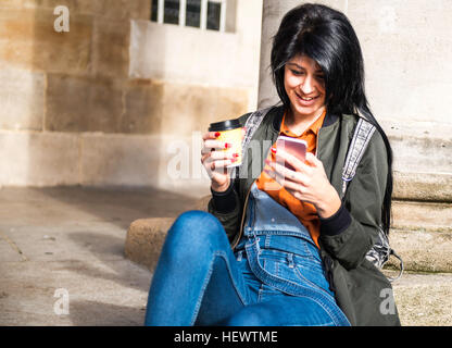 Young woman sitting beside pillar, holding takeaway coffee cup, looking at smartphone Stock Photo