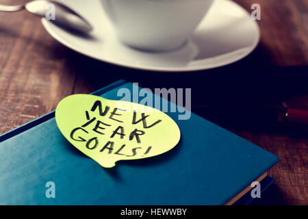 a yellow sticky note in the shape of a speech balloon with the text new years goals written in it attached to a black notepad on a rustic wooden table Stock Photo